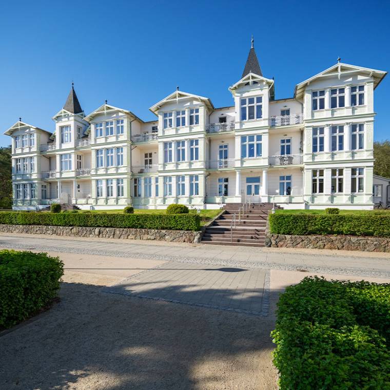 Die Strandvilla Wald & See mit weißer Fassade, Balkonen und Türmen, eingebettet in grüner Umgebung unter klarem Himmel.