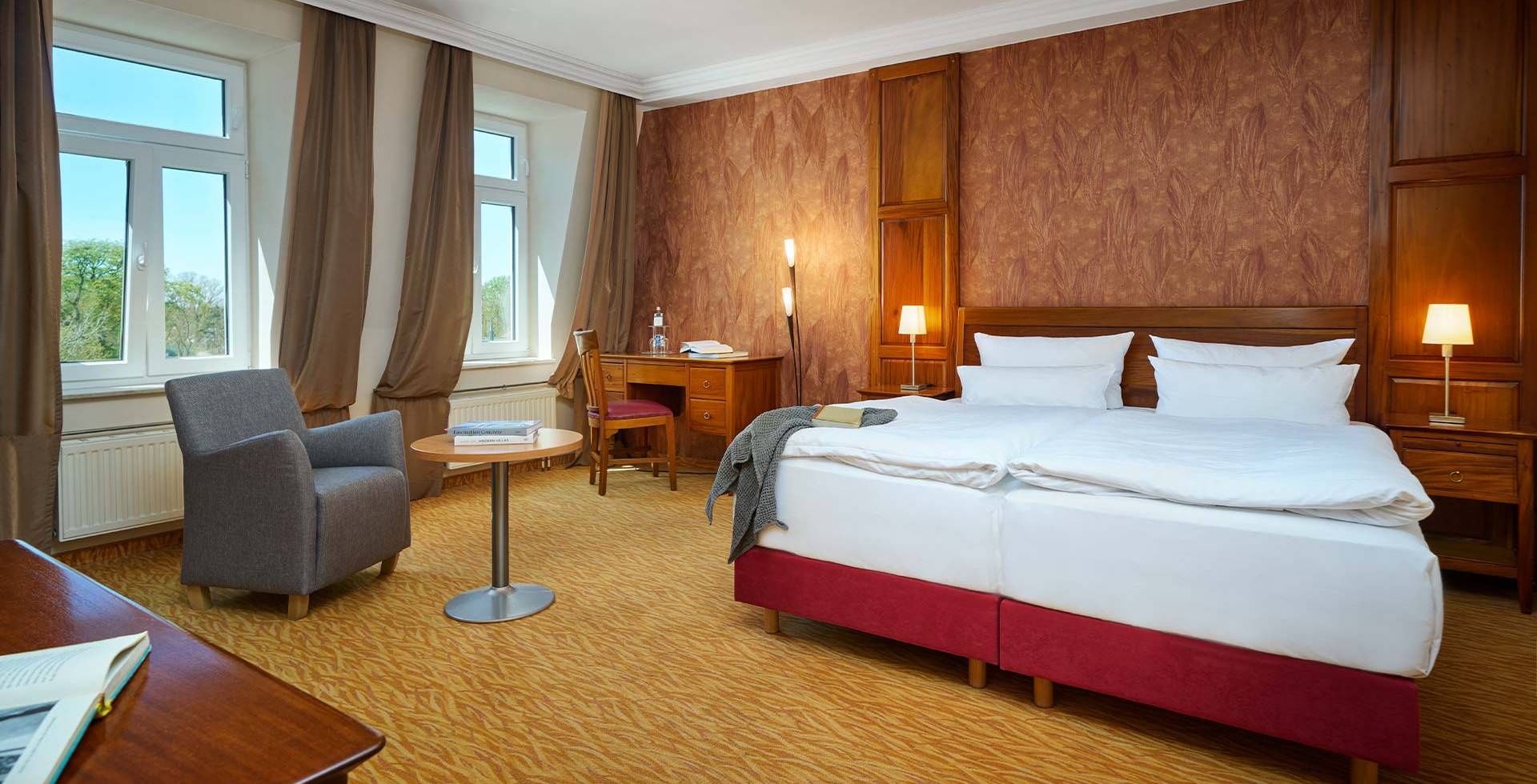 Helles Doppelzimmer im Strandhotel Preußenhof mit Sesseln, Schreibtisch, Fensterfront und Teppichboden.