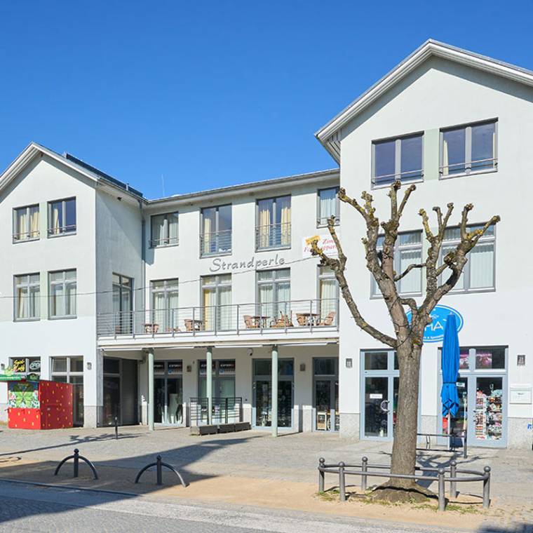 Haus Strandperle mit Souvenirständen und Ferienwohnungen, gelegen an einer belebten Promenade unter blauem Himmel.