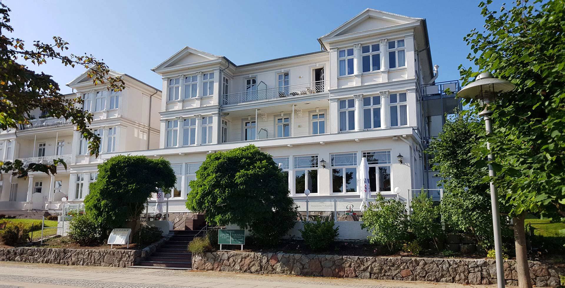 Zinnowitzer Hof mit weißer Fassade und großen Fenstern, umgeben von Bäumen und Grünflächen bei sonnigem Wetter.