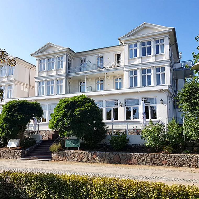 Zinnowitzer Hof mit weißer Fassade und großen Fenstern, umgeben von Bäumen und grünem Garten bei sonnigem Wetter.