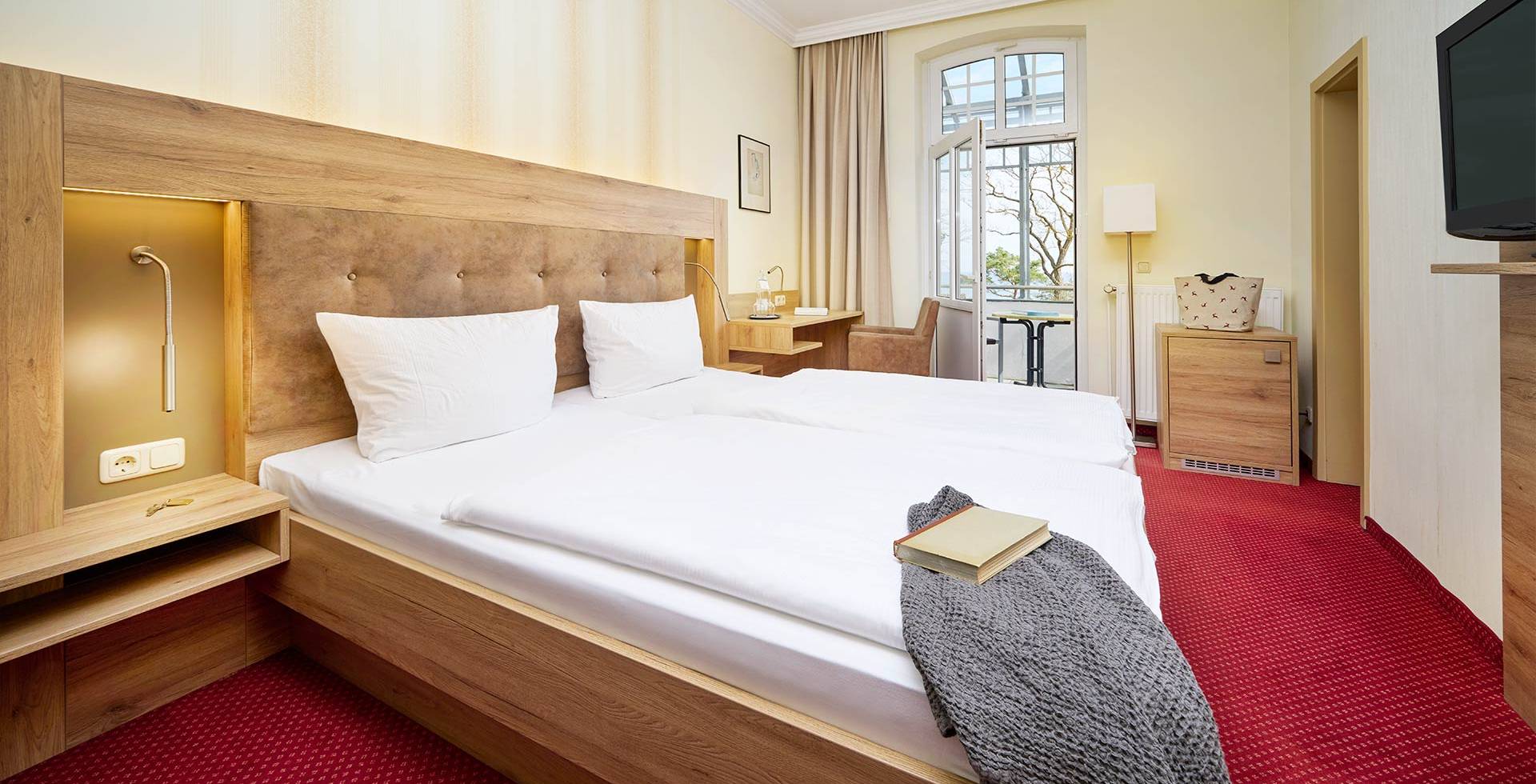 Doppelzimmer mit Bett, TV, Schreibtisch, Sessel und Fenster mit Ostseeblick.