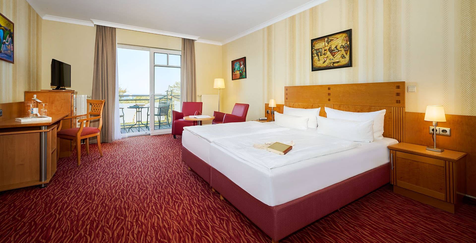 Porträt eines Doppelzimmers Meerseite im Strandhotel Prinz Preussen mit Schreibtisch, Doppelbett, roten Sesseln und Meerblick.