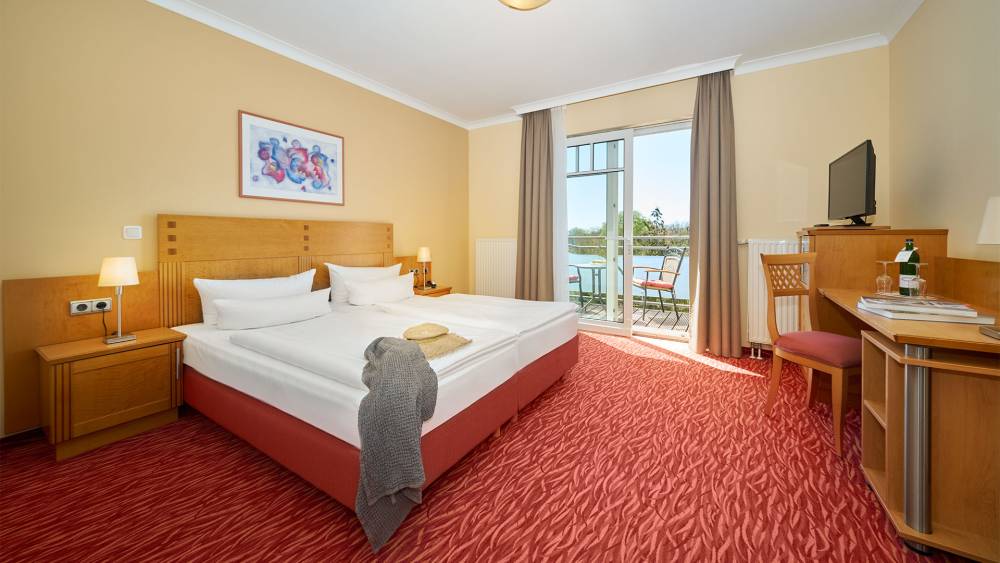 Hotelzimmers mit Doppelbett, Schreibtisch, Balkon, Sesseln und Ausblick auf die Landseite im Strandhotel Prinze von Preussen.
