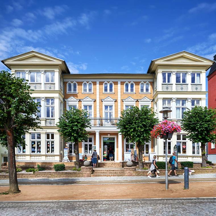 Strandvilla Viktoria mit eleganter Architektur, Bäumen und Geschäften, Spaziergänger vor der Fassade bei sonnigem Wetter.