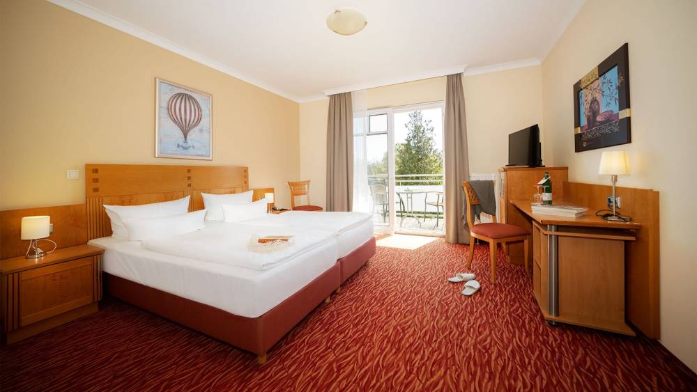 Porträt eines Doppelzimmers im Strandhotel Prinz von Preussen mit großem Bett, rotem Teppich, Minibar, TV und Sitzbereich.