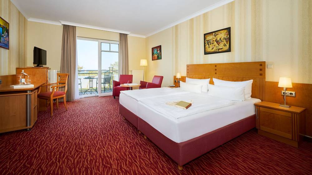 Porträt eines Doppelzimmers Meerseite im Strandhotel Prinz Preussen mit Schreibtisch, Doppelbett, roten Sesseln und Meerblick.