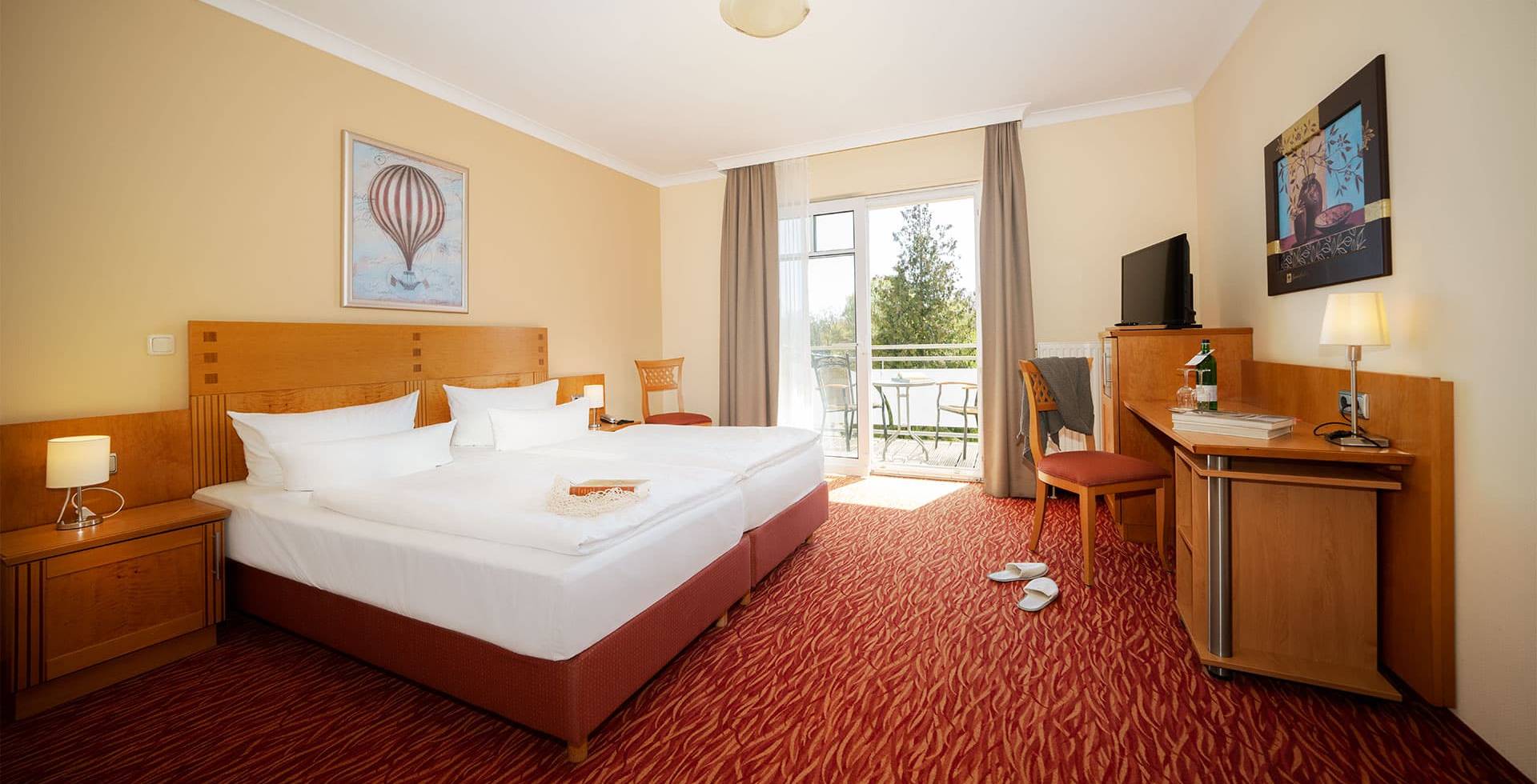 Porträt eines Doppelzimmers im Strandhotel Prinz von Preussen mit großem Bett, rotem Teppich, Minibar, TV und Sitzbereich.