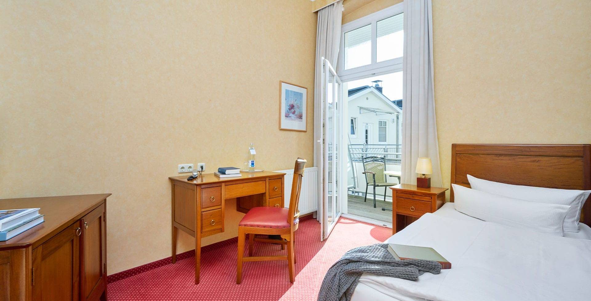Einzelzimmer Landseite in der Strandvilla Viktoria mit klassischem Holzmobiliar, Tageslicht und Zugang zum Balkon.