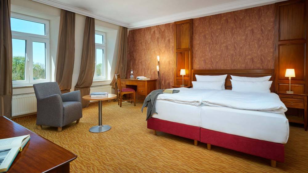 Helles Doppelzimmer im Strandhotel Preußenhof mit Sesseln, Schreibtisch, Fensterfront und Teppichboden.