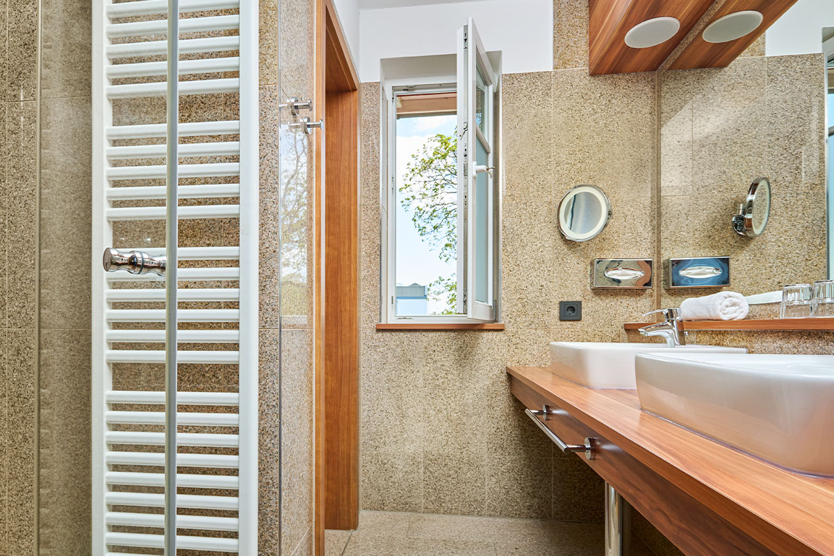Modernes Badezimmer mit Fenster, Doppelwaschbecken, Spiegeln und Handtuchwärmer im Strandhotel Kormoran.