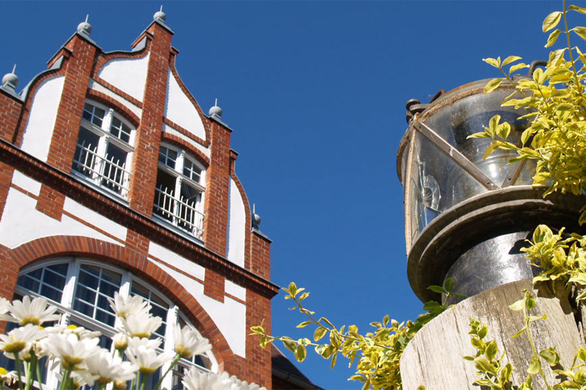 Fassade des Strandhotels Kormoran mit roten Backsteinen, Blumen und einer alten Laterne vor klarem Himmel.