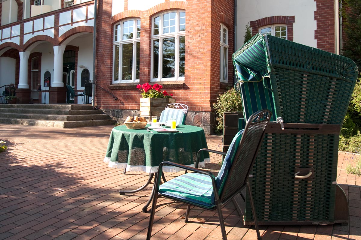 Ein gedeckter Tisch mit Frühstück auf der Terrasse des Strandhotels neben einem grünen Strandkorb bei sonnigem Wetter.