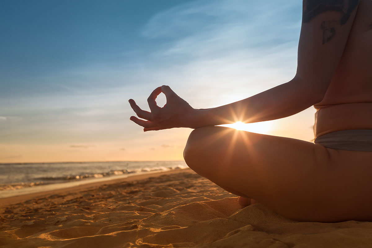 Nahaufnahme einer Person beim Meditieren am Strand, die Sonne strahlt durch die Finger bei einem ruhigen Sonnenuntergang.