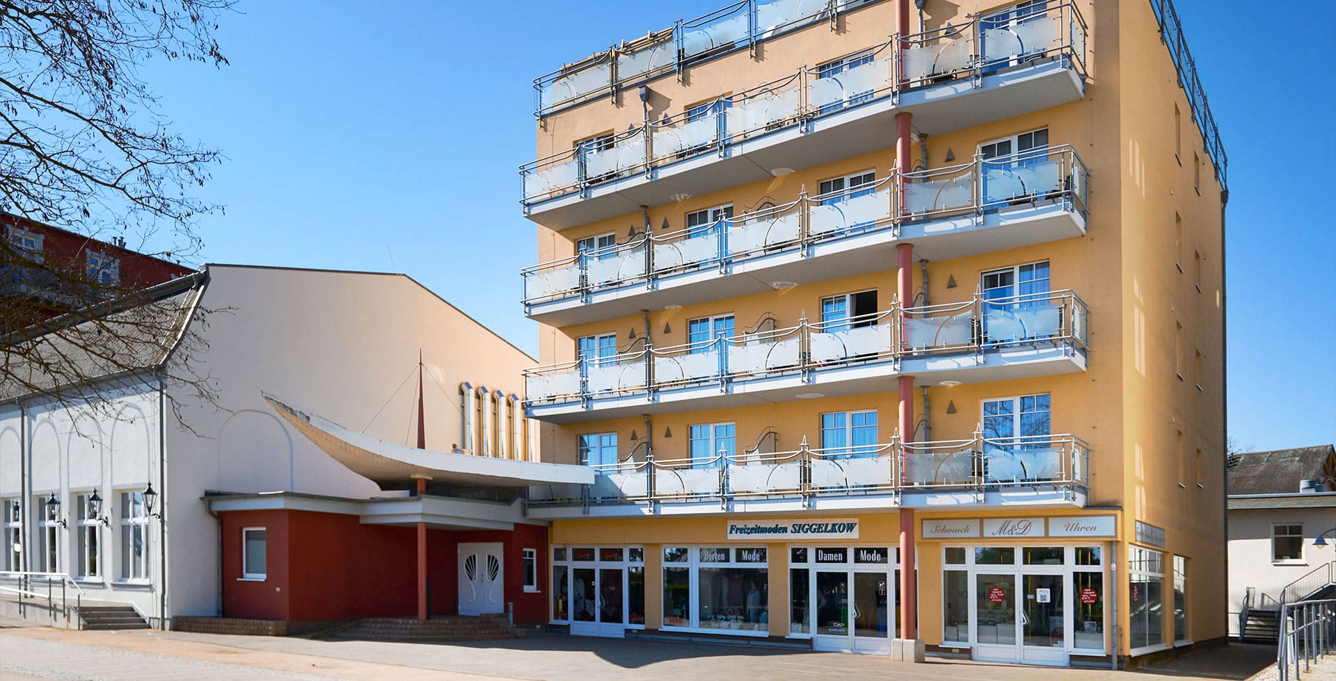 Gelbes Strandhotel Prinz von Preußen mit mehreren Balkonen und darunterliegenden Geschäften an einer sonnigen Straße.